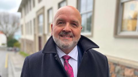A man called Jonathan Le Tocq smiles at the camera while he stands outside a building. He is bald and has a short grey beard. He is wearing a black coat with its collar pushed up. Under the coat, he is wearing a navy suit, white shirt and a pink tie with a floral pattern on it.