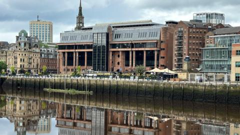 Newcastle Crown Court reflected in the River Tyne running in front of it. It is an imposing building made from smooth red stone with massive black windows and tall columns along its frontage.