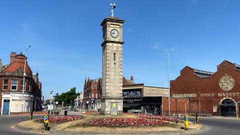 A tall, thin, grey stone clock tower on a grassy base with flower bed, with buildings including the town's market hall in the background.