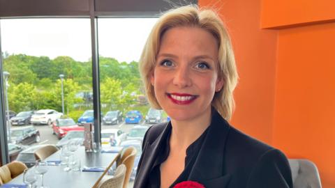 Woman with short blonde hair red lipstick black blazer stood infront of window 
