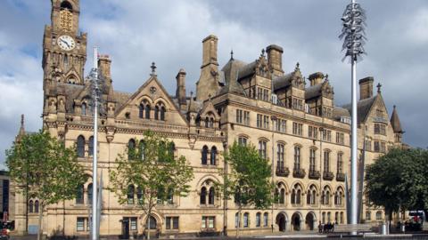 A historic city hall building with a clock tower. It sits in a tree lined square and there are benches also visible.