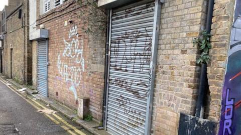 A graffiti-covered wall. There are two metal roller shutters and a black drain pipe which has a plant growing from it. There is a road which has some litter and double yellow lines. 