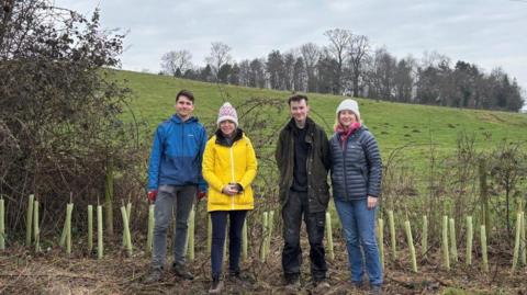 Four people stood next to each other in front of a row of juvenile trees and a green field