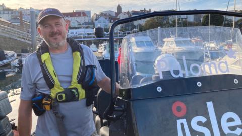 Dave Herschel stood next to his boat with a baseball cap on and a yellow and black life jacket harness. His boat has the word Paloma written on it. The boat is in a harbour surrounded by other vessels.