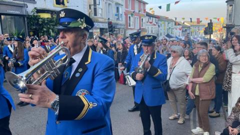 A brass band in bright blue uniforms marches through a crowded town street decorated with colourful bunting, as spectators line both sides of the road.