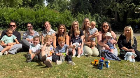 Eight women sitting in a park and smiling at the camera on a sunny day. In front of them are eight boys who are sitting down on the grass or on their mum's knee. Some are eating, others are looking at a bubble machine in the distance. Children's toys can be seen.