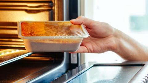 A hand puts a ready meal in a plastic container into an oven