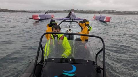 The photo shows a Guernsey Ports rescue boat towing a large racing trimaran that has capsized in open water. The trimaran is upside down, with its three hulls visible and the main structure partially submerged. Two crew members in bright yellow waterproof gear are on the rescue boat. The water is relatively calm, and the sky is overcast. The trimaran’s hulls are coloured red and purple.