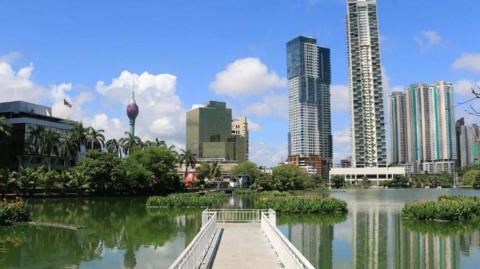 A walkway extends across a lake, surrounded by water and shrubbery. In the distance are some high-rise buildings and palm trees surrounding it. The sun is shining.