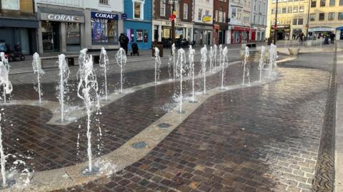 A large number of water shoots, coming out the ground in a town square. There are buildings all around, with shoppers in the area. The floor is brick. 