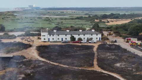A drone image of a lone row of white cottages surrounded by heathland. The area in front of the homes has been scorched due to a wildfire. There are some fire engines parked nearby. A nuclear power station is in the distance on a coastline.