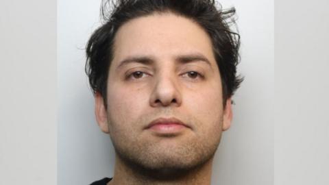 A mugshot of a man with medium-length, dark brown hair. He has short dark stubble and is looking directly into the camera.