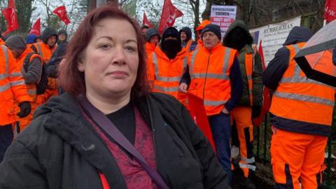 A woman with short red hair, who is wearing a black coat and purple cross-body bag is standing at a protest, surrounded by men all wearing orange, hi-vis trousers and coats.