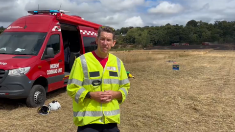 A man standing in front of a burnt field in a hi vis jacket with a red fire van behind him