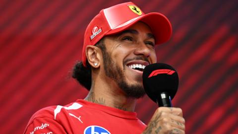 Lewis Hamilton smiling and speaking into an F1-branded microphone while wearing Ferrari kit before Hungarian Grand Prix final practice