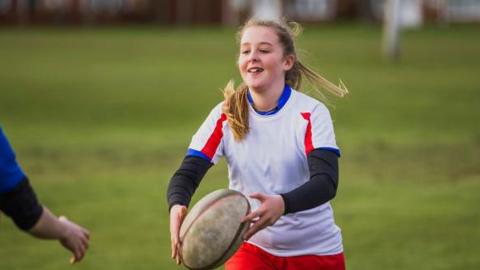 Teenage girls play rugby on a field. The girl with the ball is wearing a white T-shirt with red stripes and a blue collar over a black long-sleeve top.