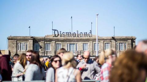 The exterior façade of the Tropicana lido on Weston-super-Mare's seafront, where the attraction was installed. Above the building there is a large sign which says Dismaland. In the out-of-focus foreground there are visitors queuing between metal fencing. 
