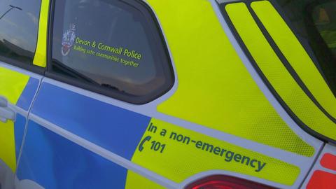 The side of a Devon and Cornwall Police car. The car is bright yellow and blue and has 'Devon and Cornwall Police Building safer communities together' written on the rear window.