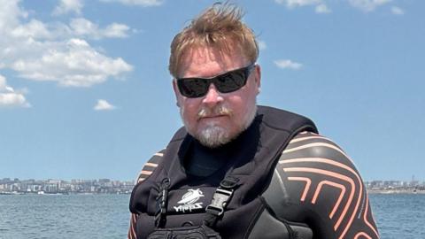 Karl Bushby, wearing sunglasses and a wetsuit, with a calm Caspian Sea in the background on a brightly sunny day. 