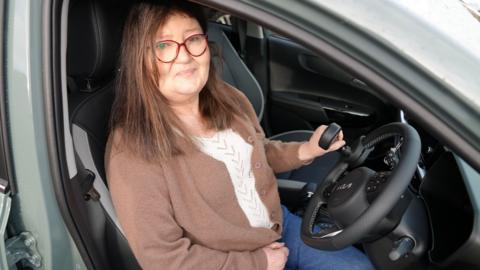 Andrea, who has brown long hair and wears a brown cardigan, sitting in a car and smiling