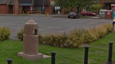 A street view image of the drinking fountain which is sitting on grass with a car park in the background