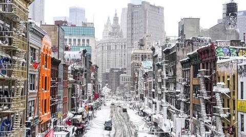 A snow covered main street lined with colourful buildings that have fire-escape stairs on the outside. In the distance are skyscrapers.