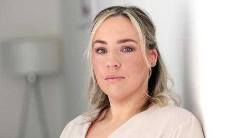 A head and shoulders picture of Maeve Bradley looking directly at the camera. She has shoulder-length hair with blonde highlights which is tied back. She is wearing a beige v-neck top or jacket.