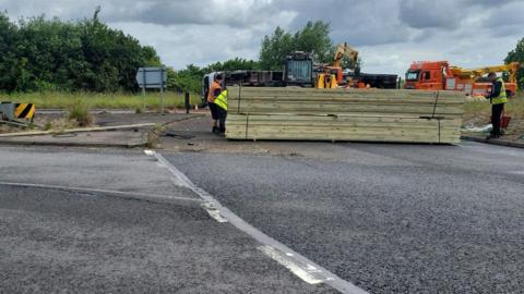 Overturned lorry on the road- with wooden planks in front of the lorry and two men in High-visibility jacket standing on either side of the wooden planks 