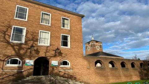 A picture of the entrance to the devon county council office. It is a large brick building with grass and trees surrounding it. 