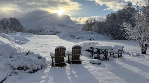 A snowy scene in Fachwen, Gwynedd. Garden furniture, covered in snow, looks out to a snowy landscape, with mountains in the background.