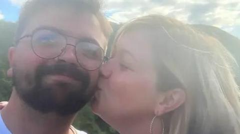 Selfie-style image of a man with a beard and wearing glasses being kissed by a woman with blonde hair.