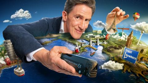Rob Brydon holding a 'DESTINATION X' bus over a map of Europe. He's also holding a small Eiffel Tower