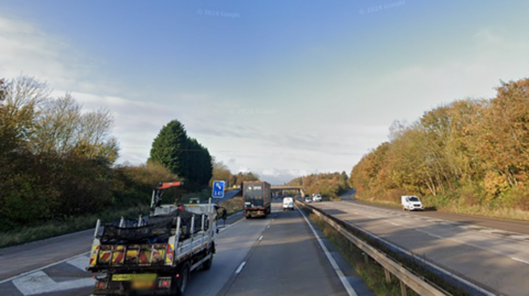 Vehicles travelling on a motorway that has two lanes. A slip road can also be seen on the far left side. There are trees lining the side of the carriageways and a concrete barrier separates the two sides of the road. The sky in the background is blue.  