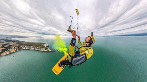 A kite surfer hundreds of feet up in the air. The photos is taken from above his left shoulder, showing the coast of the Isle of Wight below