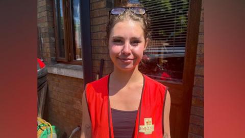 A young woman is standing beside the back door of a house. She is wearing a red high visibility vest with Royal Mail branding on it.