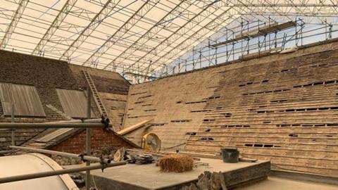 A tent over a slated roof. The roof have several tiles removed. There is also a ladder one of the roof areas.