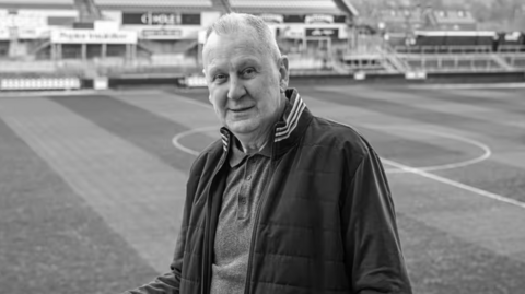 A black and white image of John with a football pitch behind him. He is smiling to camera and wearing a casual polo shirt and tracksuit top.