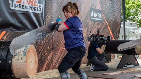 A woman with brown hair tied up, wearing a dark blue and red T-shirt and black trousers, is holding a large saw which she is using to chop through a large log of wood. There is a wooden wall in the background and signs reading "Stihl Timbersports" and "TikTok" a cameraman is in the background filming another competitor.