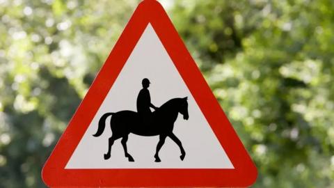 A horse and rider triangular warning road sign, with a hedge behind 