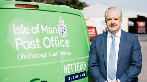 Simon Kneen standing next to a green van that reads Isle of Man Post Office, Net Zero Isle of Man. He has grey hair and is wearing a blue suit and tie with a white shirt.