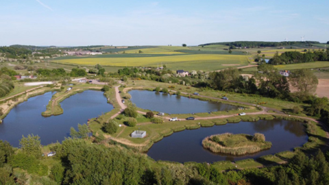 An aerial photograph taken of green fields with hedges and trees under a blue sky. Three small lakes are in the centre of the picture with stretches of land linking them. A roadway with a car on it runs around the centre lake. 