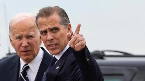 US President Joe Biden stands with his son Hunter Biden, who is pointing a finger at something off frame. 