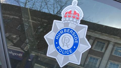 A close-up of a Humberside police badge on the window of one of their vehicles. It is a grey star, with a blue circle in the centre and is topped with a grey and red crown.
