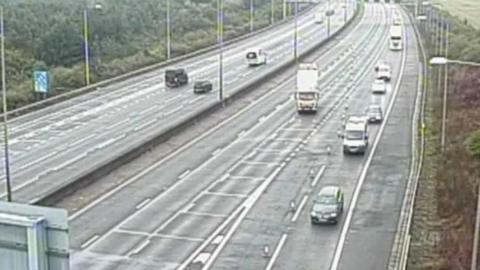 A motorway showing both directions, with a number of cars driving towards the camera and a few driving away from the camera. On either side of the road are trees. 