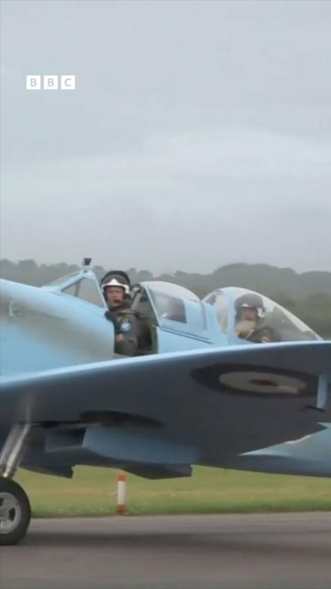 A man in a cockpit of a Spitfire on a runway, the sky is covered in grey cloud