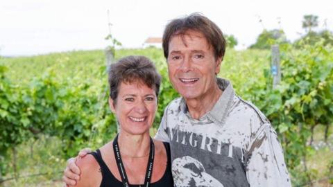 A woman with short brown hair smiles at the camera while standing next to Cliff Richard, who also smiles and has his hand on her shoulder. They stand in front on a vinyard.
