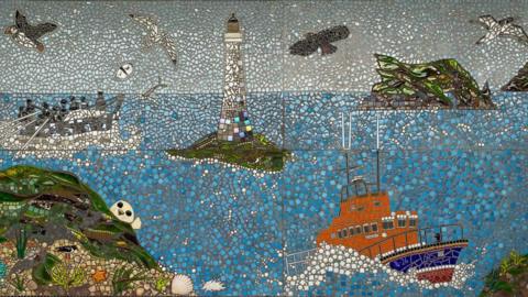 A colourful mosaic which features an old rowing boat lifeboat with men using oars, the current modern Port St Mary lifeboat, the Drinking Dragon rock formation off the southern coast, Chicken Rock lighthouse, and wildlife including a seal, gulls, puffins and a scallop shell.