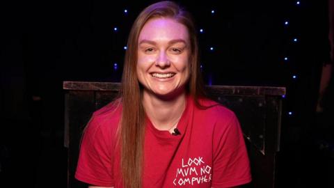 Jodie Bartle is sitting in a dark room which has blue fairy lights hanging on the wall and a table. Bartle has long hair and she is wearing a red T-shirt.