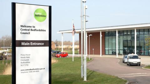 A picture of Central Bedfordshire Council Headquarters at Chicksands. The building, a modern glass-fronted construction, is in the background on the right hand side. On the left, in the foreground, is a sign saying "Welcome to Central Bedfordshire Council".