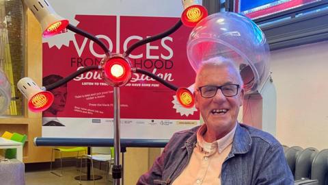 George Harris sitting under an old-fashioned helmet-style hairdryer next to a heated element with four arms with heat lamps on the ends in from of a red sign that says 'voices from the hood'